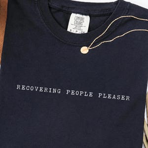 Puede incluir: Una camiseta de manga corta en color azul marino oscuro con el texto blanco RECOVERING PEOPLE PLEASER impreso en el pecho. La camiseta tiene una etiqueta de la marca Comfort Colors en el cuello interior que indica la talla S y una cadena dorada.
