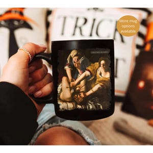 May include: Black ceramic mug featuring a classical painting with the text "GIRLS WILL BE GIRLS." The artwork depicts a dramatic scene with figures in historical attire. A small, round sticker on the right says "More mug options available."