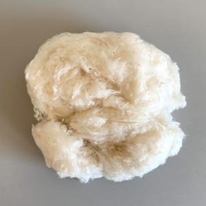 May include: A close-up of a mass of raw, unbleached cotton fibers. The fibers are a creamy off-white color with some light brown flecks, and have a soft, fluffy texture. The cotton appears to be unprocessed and ready for spinning or other textile uses.