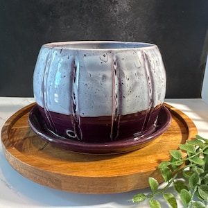 May include: A round ceramic planter with a glossy finish, featuring a two-tone design with a speckled light gray upper section and a solid deep purple lower section. The planter has vertical ribbed detailing and sits on a matching purple saucer.