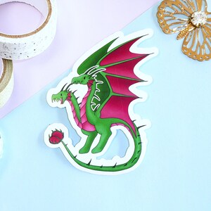 May include: A green two-headed dragon sticker with pink wings. The dragon has a long, spiky tail and is standing on its hind legs.