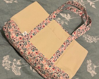 May include: A beige tote bag with floral patterned trim and handles. The trim and handles feature a design with pink, white, and green flowers on a dark background. The bag is rectangular with a simple design.