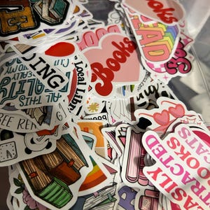 May include: A collection of colorful stickers with book-related themes. The stickers feature various designs, including stacks of books, hearts with the word "Books," and the word "READ" in rainbow colors. Other stickers have phrases like "Local Library" and "Easily Attracted to Books Cats."