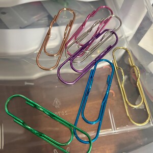 K&ouml;nnte beinhalten: Eine Sammlung farbenfroher B&uuml;roklammern in verschiedenen metallischen Ausf&uuml;hrungen, darunter Gold, Ros&eacute;gold, Lila, Blau und Gr&uuml;n. Die B&uuml;roklammern sind auf einer transparenten Kunststoffoberfl&auml;che angeordnet und zeigen ihre unterschiedlichen Farbt&ouml;ne und Formen.