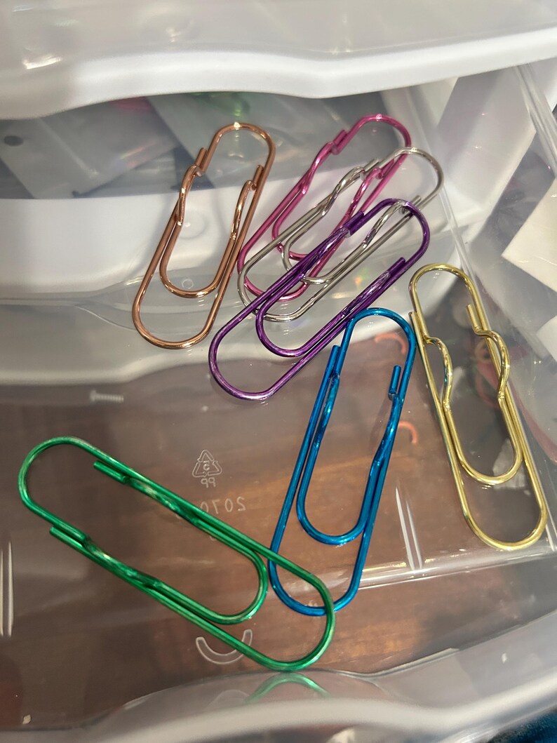 K&ouml;nnte beinhalten: Eine Sammlung farbenfroher B&uuml;roklammern in verschiedenen metallischen Ausf&uuml;hrungen, darunter Gold, Ros&eacute;gold, Lila, Blau und Gr&uuml;n. Die B&uuml;roklammern sind auf einer transparenten Kunststoffoberfl&auml;che angeordnet und zeigen ihre unterschiedlichen Farbt&ouml;ne und Formen.