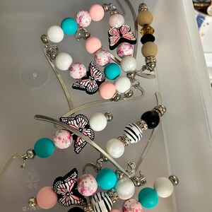 K&ouml;nnte beinhalten: Eine Sammlung von Perlen-Lesezeichen in einem transparenten Kunststoffbeh&auml;lter. Die Lesezeichen haben Perlen in Rosa, Wei&szlig;, T&uuml;rkis und Schwarz, mit Schmetterlings- und Zebra-Akzenten. Die Metallarme der Lesezeichen sind silberfarben.