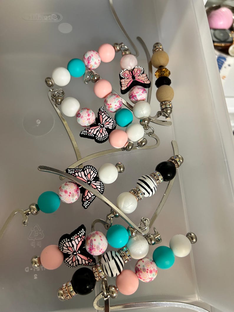 K&ouml;nnte beinhalten: Eine Sammlung von Perlen-Lesezeichen in einem transparenten Kunststoffbeh&auml;lter. Die Lesezeichen haben Perlen in Rosa, Wei&szlig;, T&uuml;rkis und Schwarz, mit Schmetterlings- und Zebra-Akzenten. Die Metallarme der Lesezeichen sind silberfarben.