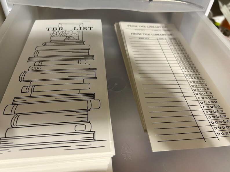 K&ouml;nnte beinhalten: Ein Stapel wei&szlig;er TBR-Listenkarten mit einer schwarzen Strichzeichnung von B&uuml;chern und dem Text "TBR LIST". Daneben ein Stapel linierter Karten mit dem Text "FROM THE LIBRARY OF" und Sternbewertungsabschnitten.