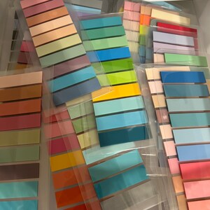 K&ouml;nnte beinhalten: Eine Sammlung farbenfroher Haftnotizbl&ouml;cke in verschiedenen Farbt&ouml;nen, darunter Rosa, Orange, Gr&uuml;n und Blau. Jeder Block enth&auml;lt mehrere rechteckige Haftnotizen, die ordentlich angeordnet und in transparenten Kunststoffh&uuml;llen verpackt sind. Die Notizen sind nach Farbfamilien geordnet.