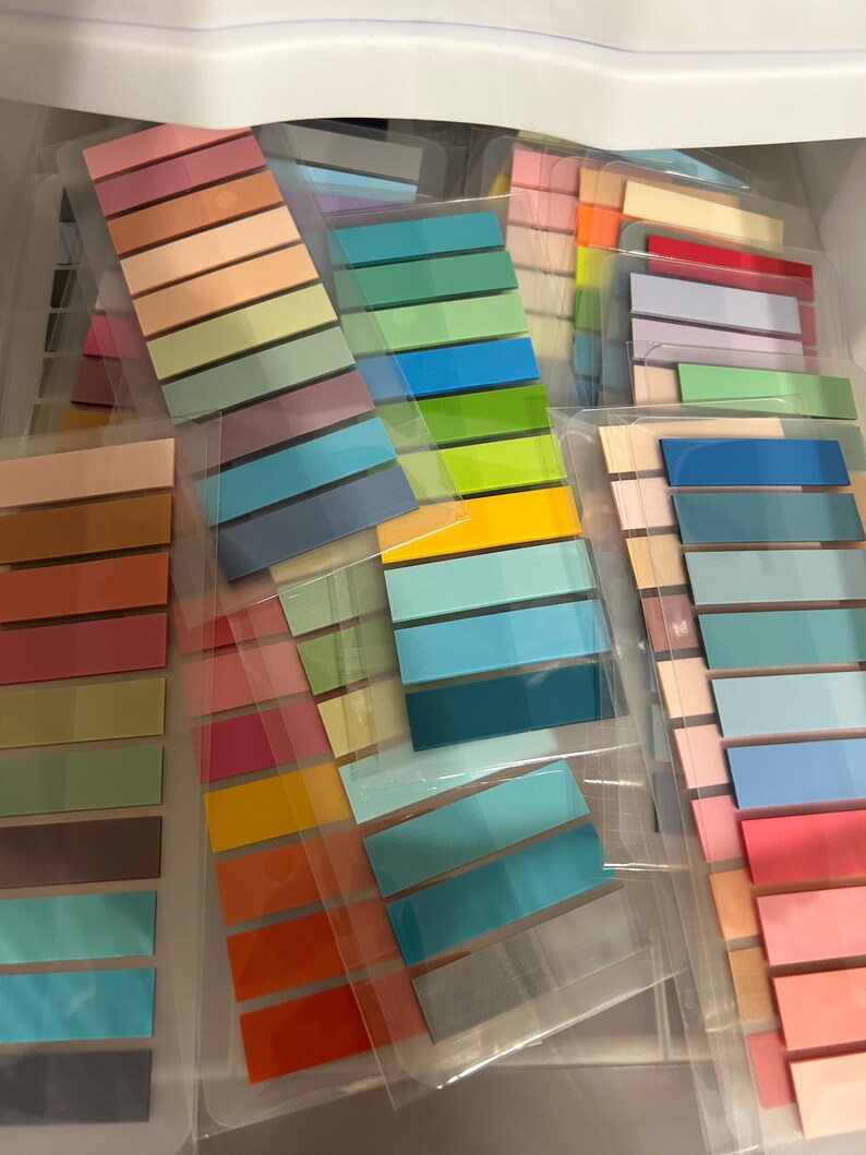 K&ouml;nnte beinhalten: Eine Sammlung farbenfroher Haftnotizbl&ouml;cke in verschiedenen Farbt&ouml;nen, darunter Rosa, Orange, Gr&uuml;n und Blau. Jeder Block enth&auml;lt mehrere rechteckige Haftnotizen, die ordentlich angeordnet und in transparenten Kunststoffh&uuml;llen verpackt sind. Die Notizen sind nach Farbfamilien geordnet.