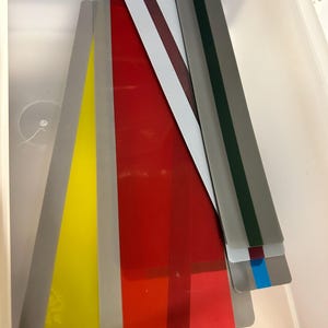 K&ouml;nnte beinhalten: Eine Sammlung farbiger, rechteckiger, transparenter Kunststoffplatten in einer wei&szlig;en Aufbewahrungsbox. Die Platten sind in verschiedenen Farben gehalten, darunter Gelb, Rot und Grau, wobei einige Streifen in verschiedenen Farben aufweisen. Die Platten sind unterschiedlich lang.