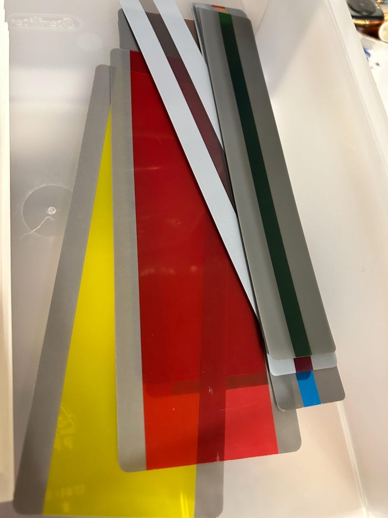 K&ouml;nnte beinhalten: Eine Sammlung farbiger, rechteckiger, transparenter Kunststoffplatten in einer wei&szlig;en Aufbewahrungsbox. Die Platten sind in verschiedenen Farben gehalten, darunter Gelb, Rot und Grau, wobei einige Streifen in verschiedenen Farben aufweisen. Die Platten sind unterschiedlich lang.