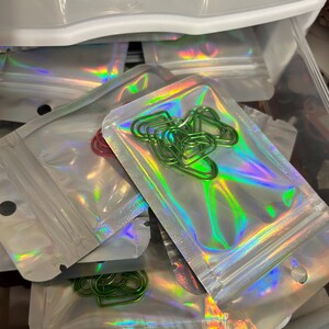 K&ouml;nnte beinhalten: Mehrere kleine, transparente, holografische Rei&szlig;verschlussbeutel gef&uuml;llt mit bunten B&uuml;roklammern. Die B&uuml;roklammern sind in verschiedenen Formen und Farben, darunter Gr&uuml;n, Rot und Blau. Die Beutel befinden sich in einer transparenten Plastikschublade.