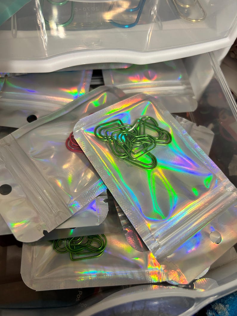 K&ouml;nnte beinhalten: Mehrere kleine, transparente, holografische Rei&szlig;verschlussbeutel gef&uuml;llt mit bunten B&uuml;roklammern. Die B&uuml;roklammern sind in verschiedenen Formen und Farben, darunter Gr&uuml;n, Rot und Blau. Die Beutel befinden sich in einer transparenten Plastikschublade.