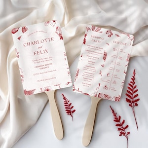 May include: Two wedding fans with wooden handles. The fans are white with burgundy text and floral illustrations. One fan lists the wedding details: names, date, time, and location. The other fan has a wedding party timeline.
