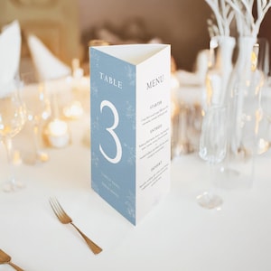 Peut inclure: Un carton de menu de table triangulaire avec un fond bleu clair et des motifs floraux blancs. Le carton affiche le numéro "3" dans une grande police, ainsi que les mots "TABLE" et "MENU". Le menu liste "STARTER", "ENTREES" et "DESSERT".