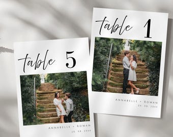 May include: Two white table number cards with black calligraphy. The cards feature a photo of a couple on a set of stone steps in a garden. The text on the cards reads "table 1" and "table 5" with the names "Annabelle & Rowan" and the date "20.08.20xx".