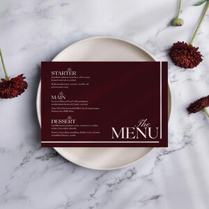 May include: A menu card with a burgundy background and white text, listing starter, main, and dessert options. The menu is on a white plate, with decorative flowers and a marble background.