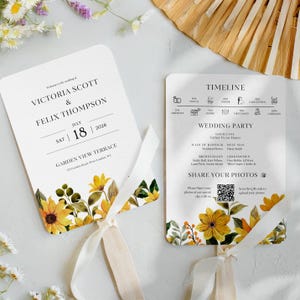 May include: A wedding program fan with a floral design featuring sunflowers and greenery. The fan is white with a cream-colored ribbon. The text on the fan reads "Victoria Scott & Felix Thompson" and "July 18, 2026".