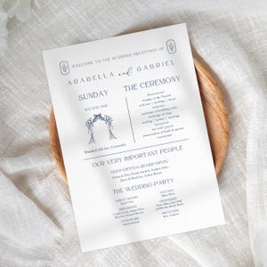 Könnte beinhalten: Ein weißes Hochzeitsprogramm mit marineblauem Text, mit den Namen Arabella und Gabriel. Das Programm enthält die Zeremoniedetails, die Hochzeitsgesellschaft und wichtige Personen. Das Programm liegt auf einem Holztablett.