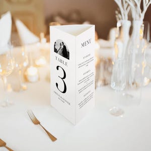 Op de afbeelding: Een witte, driehoekige tafelkaart met een zwart-wit foto van een stel. De kaart leest "TABLE 3" in grote zwarte letters, met de namen "Amelia & Oliver" en de datum eronder. De rechterkant van de kaart geeft een menu weer.