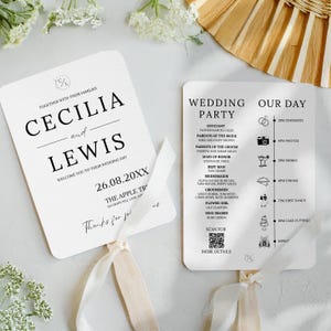 May include: Two white wedding fans with rounded edges. One fan features the names "Cecilia and Lewis" and wedding details. The other fan lists the wedding party and schedule. Both fans have cream-colored ribbon handles.