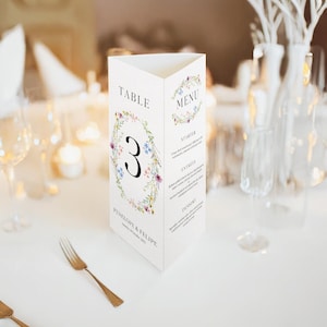 Könnte beinhalten: Eine weiße Tischnummerkarte mit einem floralen Kranzdesign. Die Nummer "3" befindet sich in der Mitte des Kranzes. Die Karte enthält auch den Text "Tisch" und "Penelope & Felipe" mit einem Hochzeitsdatum. Die Rückseite der Karte enthält ein Menü mit den Überschriften "Vorspeise", "Hauptgerichte" und "Dessert".