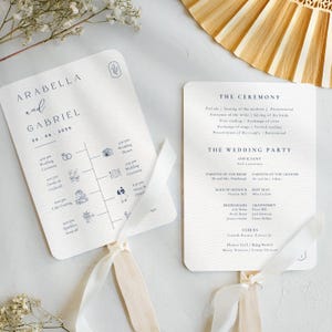 May include: A white wedding ceremony fan with a blue ribbon. The fan features a timeline of wedding events with icons and the names Arabella and Gabriel with the date 26.06.2026. The back of the fan lists the wedding party and ceremony details.