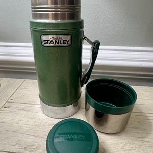 May include: Green Stanley thermos with a silver lid and base, a black handle, and a matching green lid and cup. The thermos has the "Aladdin Stanley" logo on the front. The cup and lid are separate from the thermos.