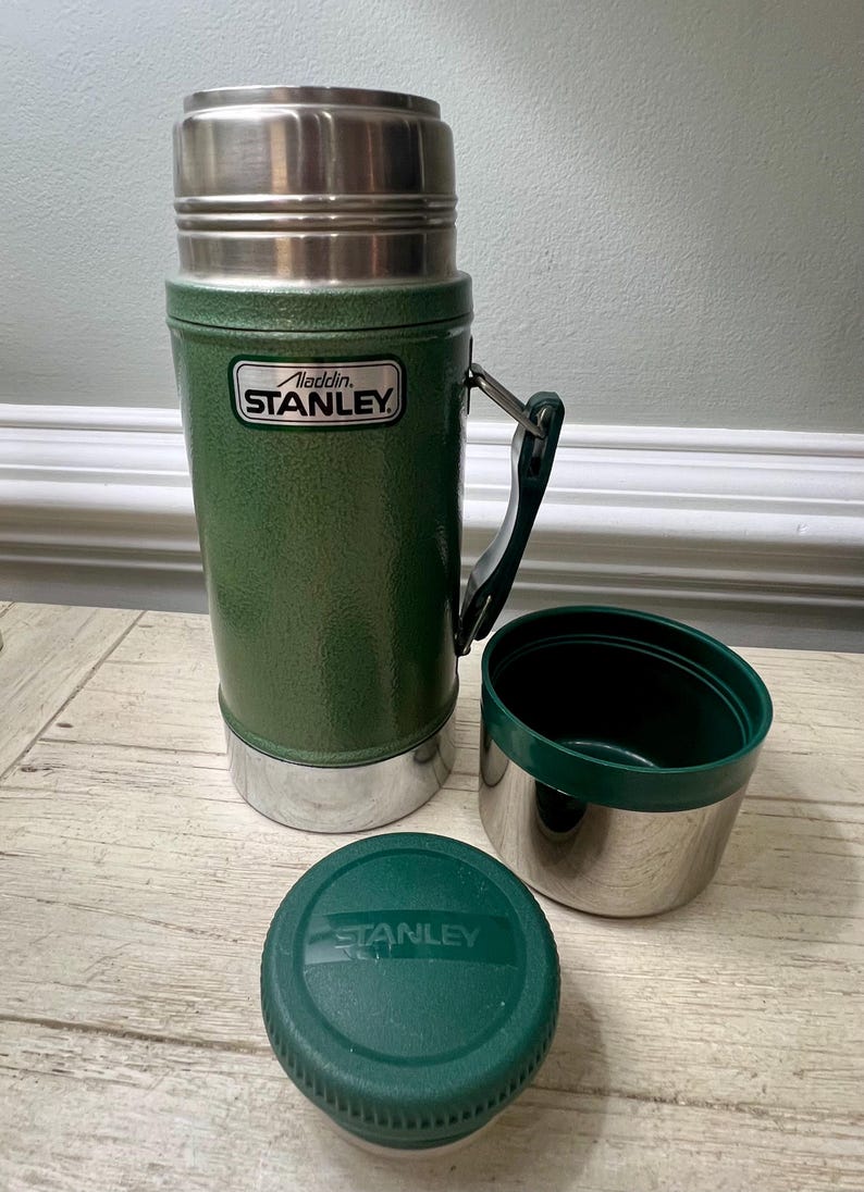 May include: Green Stanley thermos with a silver lid and base, a black handle, and a matching green lid and cup. The thermos has the "Aladdin Stanley" logo on the front. The cup and lid are separate from the thermos.