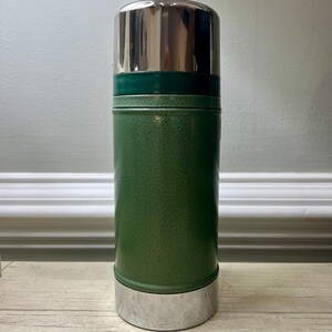 May include: A green Stanley thermos with a silver lid and base. The thermos has a textured finish and a cylindrical shape. The lid is shiny and reflective. The thermos is designed to keep beverages hot or cold.