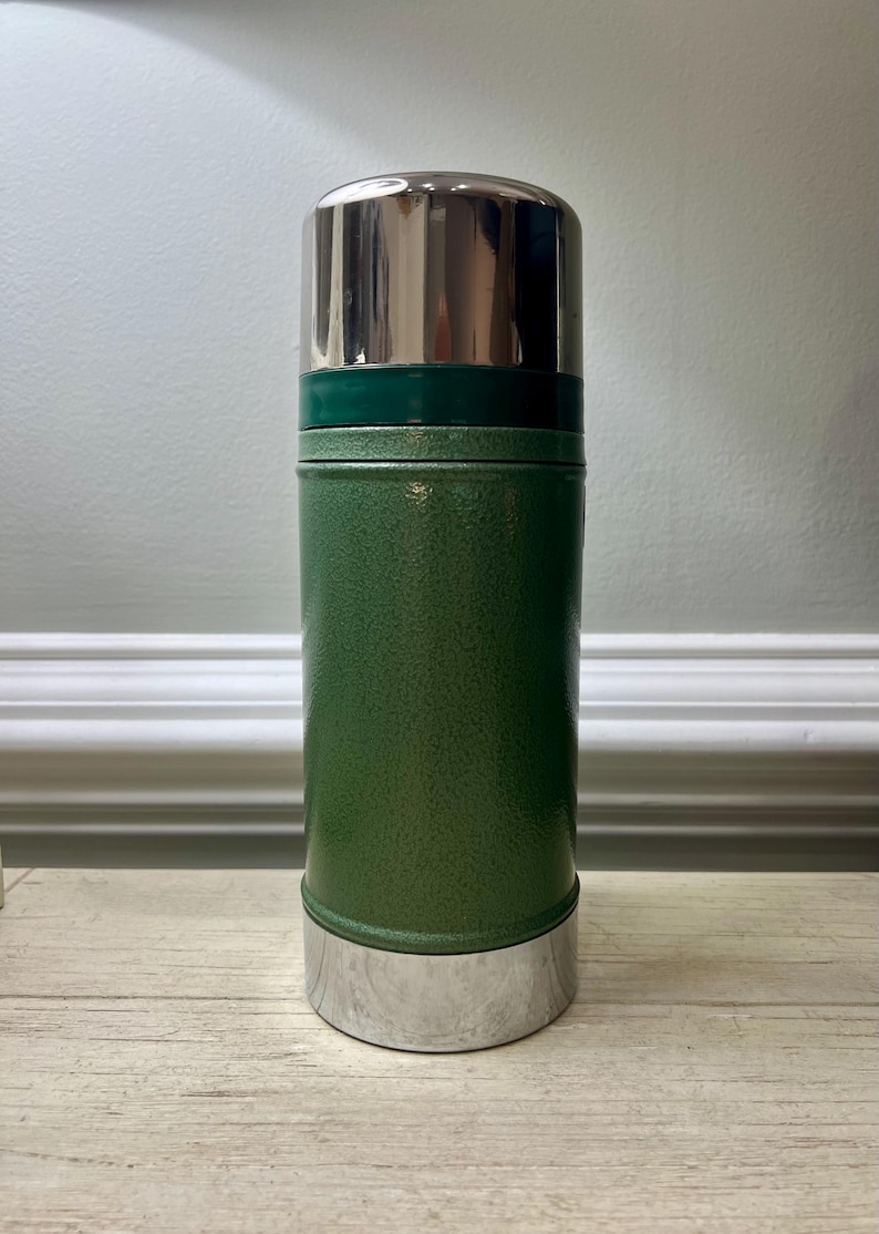 May include: A green Stanley thermos with a silver lid and base. The thermos has a textured finish and a cylindrical shape. The lid is shiny and reflective. The thermos is designed to keep beverages hot or cold.