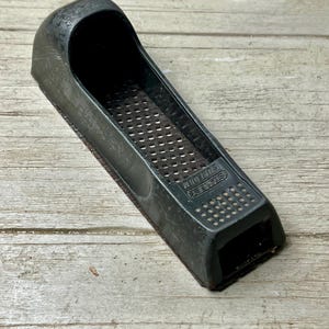 May include: A dark gray Stanley Surform tool, a hand-held rasp with a curved handle and a perforated metal surface. The tool is designed for shaping wood and other materials. The text "STANLEY SURFORM" is visible on the side.