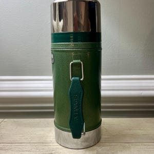 May include: Green Stanley thermos with a silver lid and base. The thermos has a handle and the word "STANLEY" is printed on the handle. The thermos is cylindrical and appears to be made of metal.