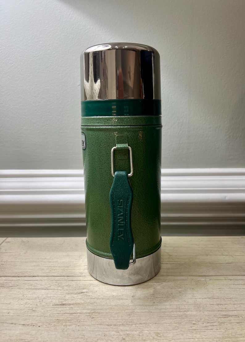 May include: Green Stanley thermos with a silver lid and base. The thermos has a handle and the word "STANLEY" is printed on the handle. The thermos is cylindrical and appears to be made of metal.