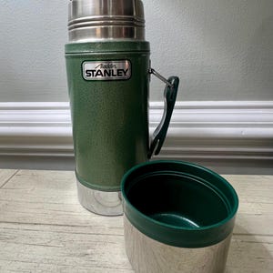 May include: Green Stanley thermos with a silver base and lid, and a matching green cup. The thermos has a handle and the Aladdin Stanley logo. The cup is detached and sits next to the thermos. The thermos is designed to keep liquids hot or cold.