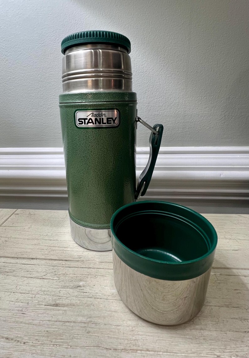 May include: Green Stanley thermos with a silver base and lid, and a matching green cup. The thermos has a handle and the Aladdin Stanley logo. The cup is detached and sits next to the thermos. The thermos is designed to keep liquids hot or cold.