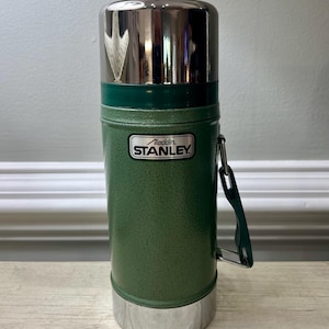 May include: Green Stanley Aladdin thermos with a shiny silver lid and base. The thermos has a green handle and a metal label that reads "Stanley". The thermos is designed to keep liquids hot or cold.