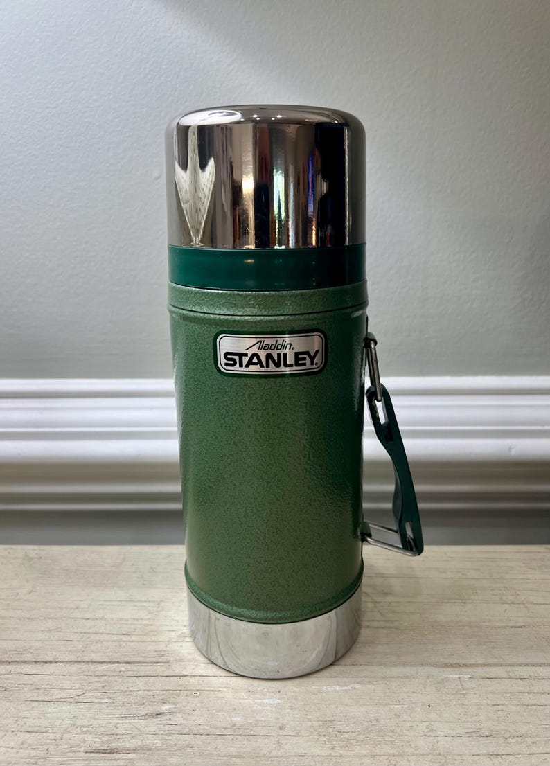 May include: Green Stanley Aladdin thermos with a shiny silver lid and base. The thermos has a green handle and a metal label that reads "Stanley". The thermos is designed to keep liquids hot or cold.