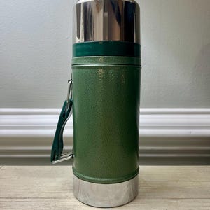 May include: Green insulated thermos with a stainless steel lid and base. The thermos has a green handle and a textured finish. It is designed to keep beverages hot or cold. A classic travel accessory.