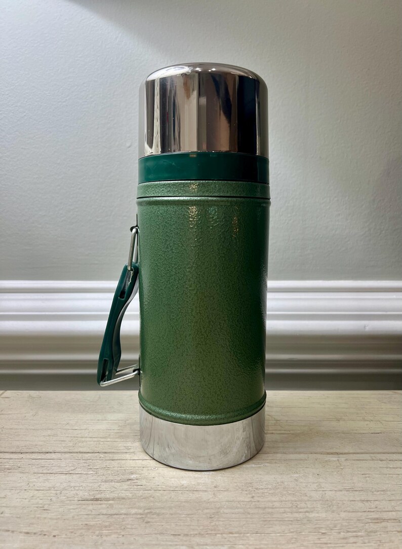 May include: Green insulated thermos with a stainless steel lid and base. The thermos has a green handle and a textured finish. It is designed to keep beverages hot or cold. A classic travel accessory.