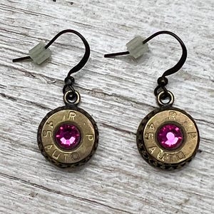 May include: A pair of handmade earrings featuring brass bullet casings with a bright pink gemstone center. The earrings have dark metal hooks and are displayed on a light wood-grain surface.