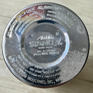 May include: Close-up of the bottom of a Stanley vacuum bottle. The silver metal surface has stamped text including "Aladdin STANLEY" and "24 oz. wide mouth". The bottle has a wide mouth and is made by Aladdin Industries, Inc.