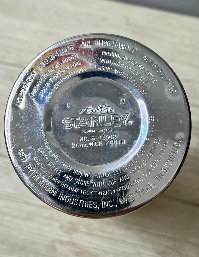 May include: Close-up of the bottom of a Stanley vacuum bottle. The silver metal surface has stamped text including "Aladdin STANLEY" and "24 oz. wide mouth". The bottle has a wide mouth and is made by Aladdin Industries, Inc.