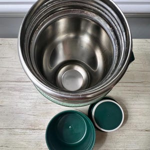 May include: A stainless steel insulated food jar with a green base and lid. The jar has a wide mouth and a green plastic bowl and lid are next to it. The interior of the jar is silver.