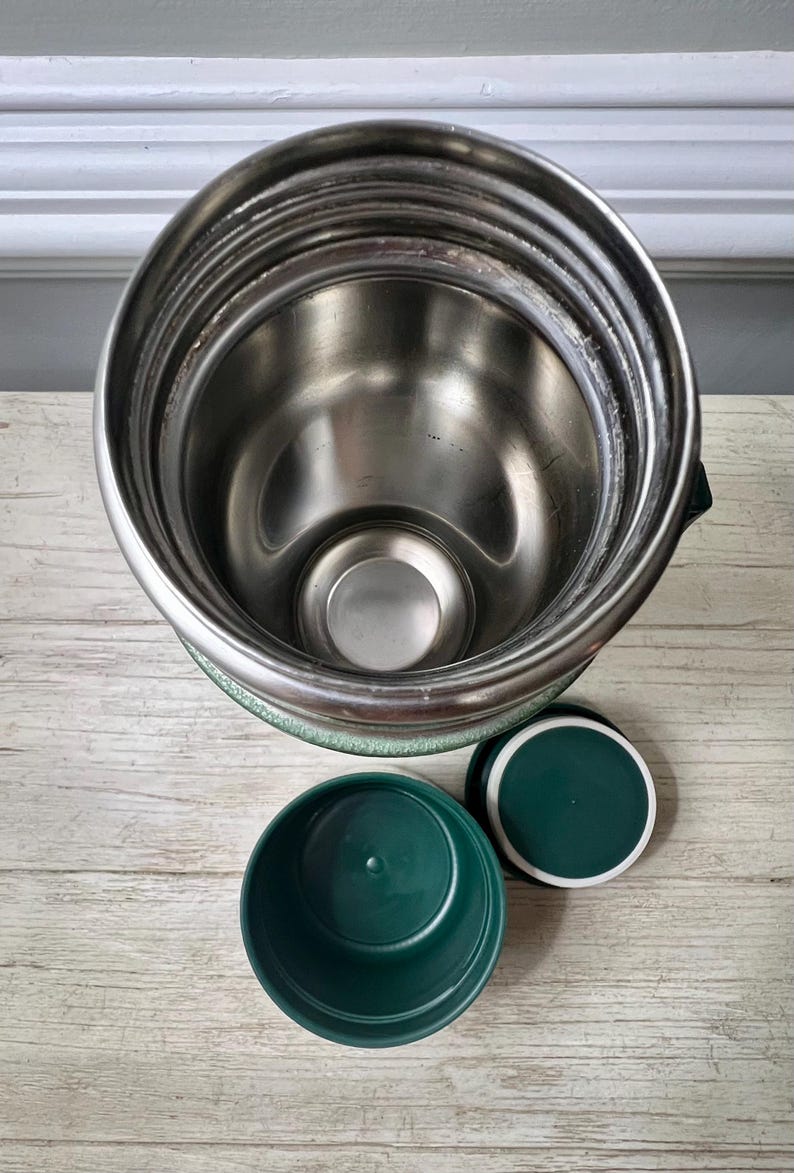 May include: A stainless steel insulated food jar with a green base and lid. The jar has a wide mouth and a green plastic bowl and lid are next to it. The interior of the jar is silver.