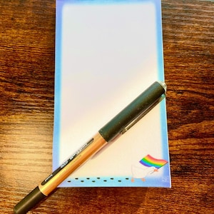May include: A blue notepad with a white goose holding a rainbow flag. The notepad has a blue border and a white interior. A black and gold pen is lying on top of the notepad.