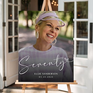 May include: A sign on a wooden easel displays a photo of a smiling woman wearing a visor and hoop earrings. The sign reads "Seventy" in script, with the name "Ellie Sanderson" and the date "05.25.2025" below.
