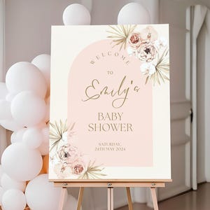 May include: Baby shower welcome sign featuring a blush pink background, gold text, and watercolor floral design. The sign reads 'Welcome to Emily's Baby Shower, Saturday, 24th May 2024'.  It's displayed on a wooden easel. The design includes palm leaves and peonies in soft peach and beige tones. The arch shape adds a modern touch to the elegant floral theme. The sign is perfect for a sophisticated baby shower celebration.