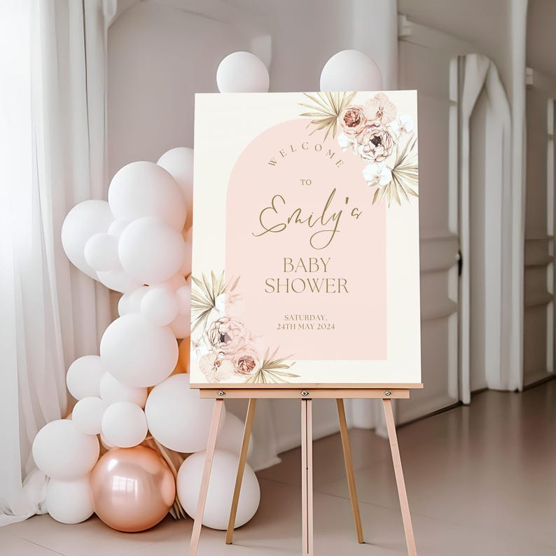 May include: Baby shower welcome sign featuring a blush pink background, gold text, and watercolor floral design. The sign reads 'Welcome to Emily's Baby Shower, Saturday, 24th May 2024'.  It's displayed on a wooden easel. The design includes palm leaves and peonies in soft peach and beige tones. The arch shape adds a modern touch to the elegant floral theme. The sign is perfect for a sophisticated baby shower celebration.